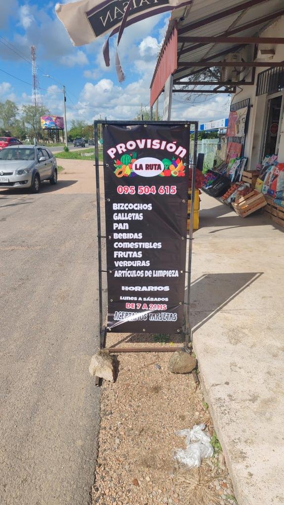 Lona impresa en estructura metálica en la vereda de un almacén de barrio, con lista de productos y horarios