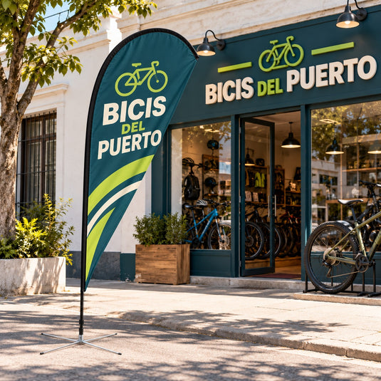 Imagen conceptual de una bandera gota de tela petróleo y lima apenas translúcida, sobre una cruceta baja de brazos metálicos finos frente a la bicicletería ficticia BICIS DEL PUERTO.