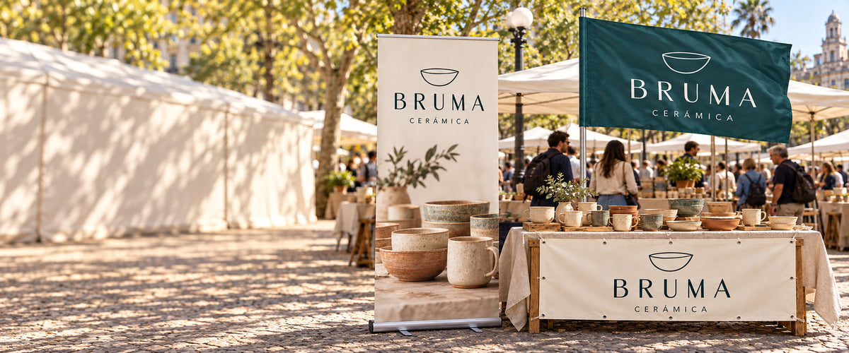 Imagen conceptual del Pack Feria BRUMA Cerámica: roll up con base de aluminio, bandera rectangular con vaina lateral en asta y lona reforzada con ojales al frente de la mesa, en una feria uruguaya. Espacio tranquilo a la izquierda para el titular de la web.