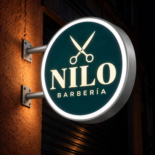 Imagen conceptual de un cartel luminoso LED circular de doble faz para NILO Barbería, con aro blanco perimetral encendido, frente azul petróleo y canto de aluminio gris satinado, montado en voladizo de noche.