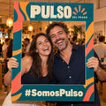 Imagen conceptual de dos personas adultas sosteniendo un marco de fotos físico y calado de la marca ficticia Pulso del Prado, con el hashtag SomosPulso impreso y el fondo real del evento visible.