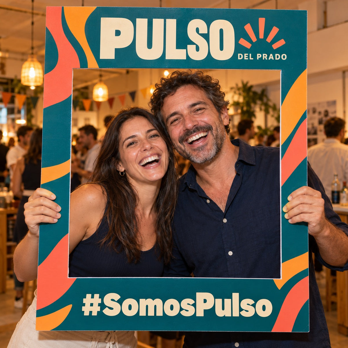 Imagen conceptual de dos personas adultas sosteniendo un marco de fotos físico y calado de la marca ficticia Pulso del Prado, con el hashtag SomosPulso impreso y el fondo real del evento visible.