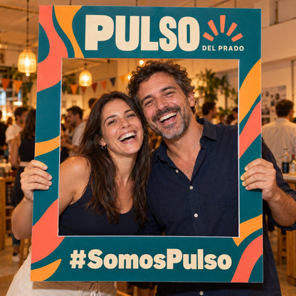 Imagen conceptual de dos personas adultas sosteniendo un marco de fotos físico y calado de la marca ficticia Pulso del Prado, con el hashtag SomosPulso impreso y el fondo real del evento visible.