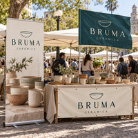 Imagen conceptual del Pack Feria BRUMA Cerámica: roll up con base de aluminio, bandera rectangular con vaina lateral en asta y lona reforzada con ojales al frente de la mesa, en una feria uruguaya.