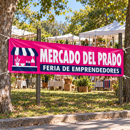 Imagen conceptual de un pasacalle magenta y azul de Mercado del Prado, atado temporalmente entre árboles junto al alambrado lateral de una feria de emprendedores.