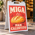 Imagen conceptual de un caballete plástico blanco abierto en la vereda de una panadería, con el afiche coral de la marca ficticia Miga y el mensaje Pan calentito impreso.